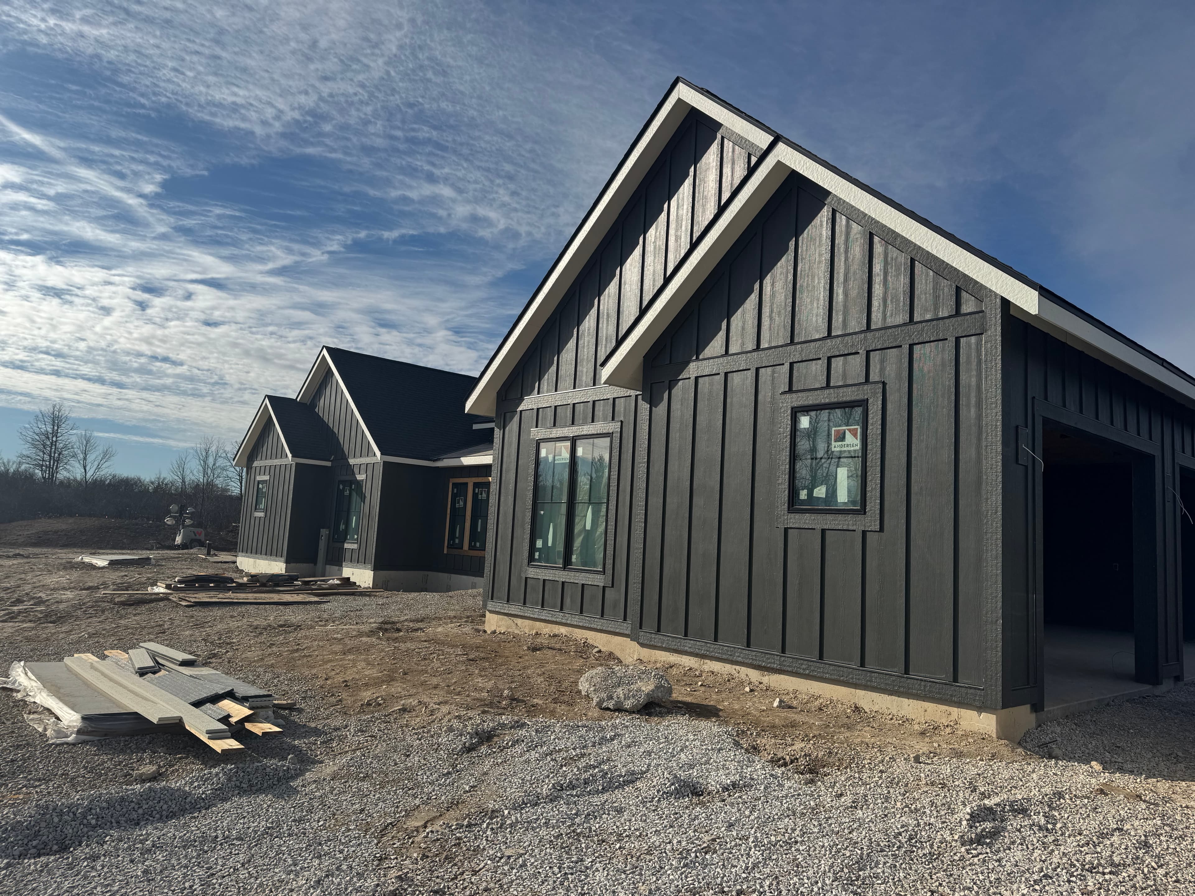Board-and-batten custom home under construction with siding staged on a gravel lot