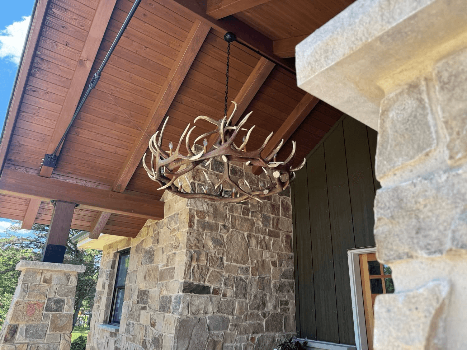Antler chandelier hung from a timber-beamed covered entry with stone columns on a custom home